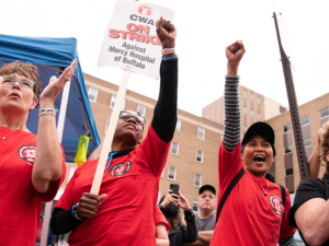 Joyful people in red shirts throw their fists in the air. One has a "CWA On Strike Against Mercy Hospital of Buffalo" picket sign. Outdoors.