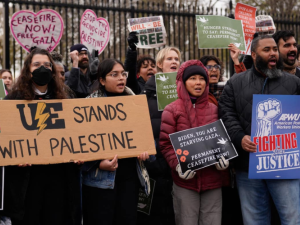 A chanting crowd stands in front of the White House fence. Three people are helping hold a large cardboard sign that says "UE stands with Palestine" with the union's lightning-bolt logo and a slice of watermelon. Other printed signs say "American Postal Workers Union: Fighting for Justice" and "Biden, you are starving Gaza. Permanent ceasefire now!" 