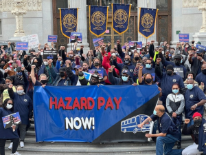 Large, multiracial, masked crowd stands on the steps of a public buliding. Many have fists in the air. Back row holds ATU banners. Front row holds a giant banner that reads "Hazard pay now!" with a line drawing of a bus.