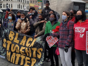 A multiracial and multigenerational crowd rallies outside. Several help hold a large banner: "BLACK STUDENTS MATTER." Others hold signs: "WE WON'T DIE FOR THE DOE" and one wears a MORE T-shirt.