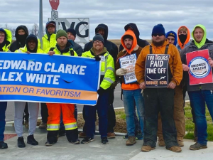 16 people stand facing the camera. Most are wearing jeans and neon yellow or orange jackets or hoodies. Several are helping to hold a blue banner: "Rehire Edward Clarke and Alex White, no retaliation or favoritism." Others hold signs: "180 hours paid time off, unionize KCVG" and "Unionize Amazon."