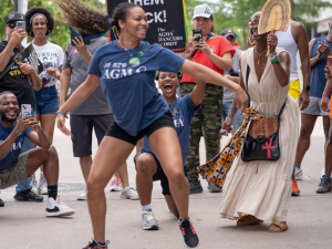 A Black dancer dances joyfully on the picket line, her long hair streaming out behind her. She is wearing a blue AGMA shirt. A delighted crowd, also mostly Black, surrounds her watching and taking photos -- some also wear AGMA shirts. The photo is full of energy, motion, and joy.