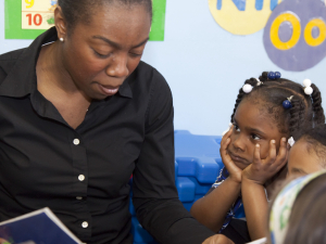 A teacher reads to kindergarteners, one of whom has her chin on her hands and seems to be rapt by the story