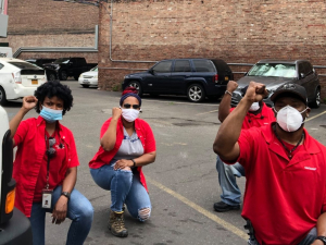 Four Black workers in red Verizon shirts kneel in a parking lot, fists raised.