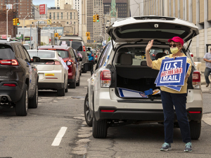 woman in front of the back of her car, trunk open, standing with a save usps sign.