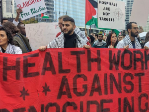 Workers stand outdoors, many wearing keffiyehs, holding a large red hand-painted banner that says "Health care workers against genocide in Gaza." Signs visible in crowd behind them say "Hospitals are not targets," "Free Palestine," and something ending in "slaughter." 