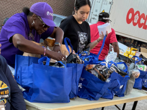 Three women work around a table with blue cloth grocery bags, outdoors. A man in a red T-shirt walks behind them. A truck is also visible.
