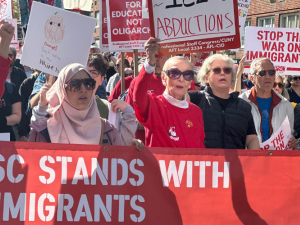 Workers, mostly women, carry a big red banner with white letters: "PSC stands with immigrants." They are in a big crowd. Some also carry picket signs, some printed and some handwritten, with messages (some partially visible) including "Dictators don't belong here," "America? Fight for it!" "Oranges don't belong in the White House," "Stop the war on immigrants," and "Stop the coup." One woman wears a hijab.