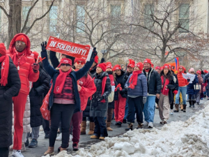 A long procession of strikers bundled up against the cold, most wearing bright red NYSNA hats or scarves, packs a sidewalk lined with piles of shoveled snow. The group is diverse in race and gender. Many are smiling. A woman in the foreground holds up a red "In Solidarity" banner.