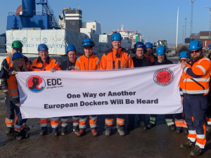 Fourteen workers in orange vests, blue hardhats, and workboots stand in a port in bright sunlight. They hold a big white banner that says "One way or another, European dockers will be heard" with logos of the SDU and the European Dockworkers Council. Most but not all appear to be white men.