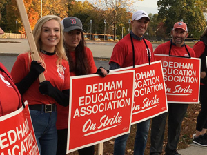 Line of workers on picket line in red union T-shirts holding signs that say "Dedham Education Association on Strike" and "Fair Contract Now" 