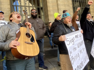 A group of people stands outside a building, singing. Edwin, a young man with guitar, has his head leaned back shouting into the sky. Several other people have fists in the air, one wears a big smile, and some hold big paper on which lyrics are written out: "Sign me up for the union, bring me my union card, We can work better with decent pay, health care, childcare, and more of a say..."