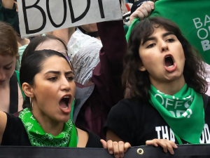 Two women in green bandanas that say “bans off our bodies” march with many others