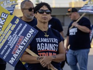 A woman with a strike picket sign and a ‘Teamsters Against Trump’ T-shirt looks past the camera