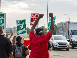 Strikers shown from behind, waving at cars, fists in air, signs read "UAW On Strike"