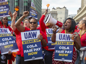 Workers in red shirts chant. Most are Black women. Many fists in air, one blowing whistle, one speaking into bullhorn. Printed signs say "UAW Stand Up, COLA and Fair Pay Now" 