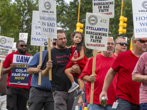 A group of people in red shirts, one man carrying a child march past the camera with signs saying Stellantis is barganing in bad faith.