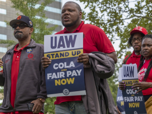 A group of workers march in Detroit holding signs that say "UAW Stand Up: Fair Pay and COLA Now." 