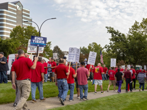 Workers in red shirts picket outside. Many of the picket signs are white with UAW logo, Local 412, and the words "Stellantis Bargaining in Bad Faith."