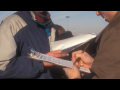 A closeup of a person’s hand signing paper on a clipboard. The person is wearing a brown uniform, possibly from UPS. Someone else is standing by in a blue and silver jacket.