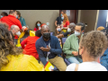 A diverse crowd of masked people talks in small groups in a conference room.