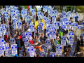 Marching crowd viewed from above, all holding printed picket signs that say "UAW on strike, unfair labor practice"