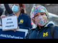 Health care workers outdoors in U of Michigan Medical Center coats. Person in front wears a colorful mask and smiles right into the camera. Behind, person holds signs "PPE OVER PROFIT" and "#HAILToTheFrontLine"