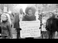 Black-and-white photo shows people marching in a New York City street, carrying signs. In the center, looking directly into the camera with a slight smile, is a Black woman with a natural hairstyle, carrying a hand-lettered sign: "Dear Black and Brown children, Your lives matter! Don't allow this corrupt system treat you otherwise! Sincerely, Your Teacher. #BlackLivesMatter"