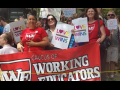 Three smiling people stand among a crowd outdoors. They hold a big red banner that says "Caucus of Working Educators" with a "WE" logo and two wear read shirts iwth the same logo. Two hold white signs with rainbow letters: "Love wins" with a heart for the letter O.  Signs carried by others behind them say "I [heart] my trans son," something ending in "LGBTQ and Trans People," and "Peace."