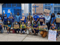 21 adults in masks outside a school. Together they hold "CAUTION" tape. Handmade signs say "100% remote start," "no mas muertes," "ready or not," "we demand safe schools," "no more martyrs for the DOE #Remote until it's safe" and "people got sick in this buidling."