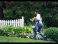 A Black woman letter carrier walks across a lawn, in front of a white picket fence, carrying the mail