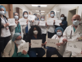 Masked health care workers in a hospital hold signs reading: "We... are... here... at... work... for.. you... Please... stay... home... for us."