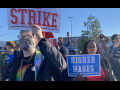 Amazon strikes hold signs saying strike, higher wages, stop racism. An Amazon facility is in the background