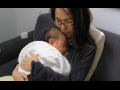 A seated woman with glasses in black holds a newborn baby wrapped in a white blanket to her chest.