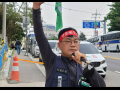 A man, Yang Hoe-dong, walks along a sidewalk wearing a resolute expression. His right arm is in the air, holding a green flag. In his left hand he holds a microphone to his lips. He wears a red headbank with printing on it in Korean, and a vest and badge that also have printed text in Korean. It seems likely he is speaking or leading chants at a union protest.