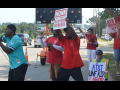Black and white men, most in red union T-shirts, picket energetically on a streetcorner. Man in front, in turquoise shirt, is talking or chanting into a mic, arm outstretched. Everyone's pose is active. Printed signs say "honk if you support workers" and "AT&T unfair, CWA on strike, ULP." One handmade sign, partially visible, says "AT&T Unfair Labor." The man in the center is holding his sign high and wind or movement blows his red shirt.