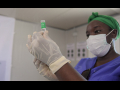 Medical officer in blue scrubs, green bandana, and surgical mask preparing vaccine for administration