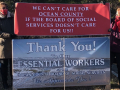 Two workers hold a banner reading “We Can't Care for Ocean County If the Board of Social Services Doesn’t Care About Us!” over the “Thank You to Our Essential Workers” sign that the Board had put up.