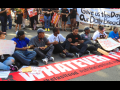 Fast food strikers, mostly young and Black, a mix of genders, sit in an intersection, linking arms. A long red banner laid out in front of them says "Whatever it takes." In the crowd behind them, some people hold a white handlettered banner: "Give us this day our daily bread."