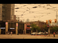 View of a city intersection. The sky, crisscrossed with power lines, is hazy grey and eerily tinted orange. Some people are out walking anyway.