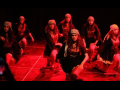 Seven women in long skirts and kerchiefs kneel, their arms in motion, apparently mid-dance. They are on a stage, bathed in red light.