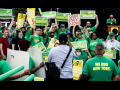 A crowd of 40 in green holds yellow and green signs saying “Fair Pay Now” and “Contract Now”