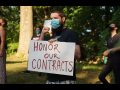 Masked person holding sign "Honor Our Contracts" in front of trees, other masked demonstrators