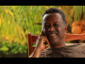 Chest-up photo of Dennis Serrette, a Black man, leaning back in a wooden rocking chair, resting his face gently on one hand, smiling warmly, bathed in warm evening light, with some greenery behind him.