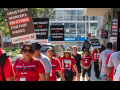 People in red T-shirts picket in the sunlight on a downtown sidewalk. Shirts say "Hotel workers strike" and "UNITE HERE Local 23." Picket signs say "Houston's workers on strike for fair wages." Another slogan partially visible on some signs is "One job should be enough."