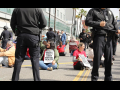 Several workers sit in the street with signs saying “Kaiser Don’t Deny Mental Health”--they are surrounded by police.