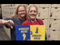Two white women stand together in front of a wall of lockers, presumably the non-public area of a grocery store, holding a printed blue and yellow sign that reads "Workers' needs vs. grocery greed," with symbols of a fist and Mr. Moneybags. The woman on the left is giving the "thumbs-down" sign. Both smile calmly.