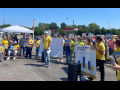 A group in a parking lot in yellow t-shirts listen while one man speaks through a microphone