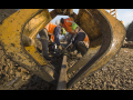 Workers in orange vests and hardhats crouch around a track. They are framed by a big yellow piece of equipment in the foreground. The day is very sunny.