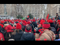 A sea of people, mostly women, of various races, bundled up in winter clothes with red hats and scarves, packs a square in New York City. One person with a megaphone stands elevated a bit in the center, surveying the crowd.